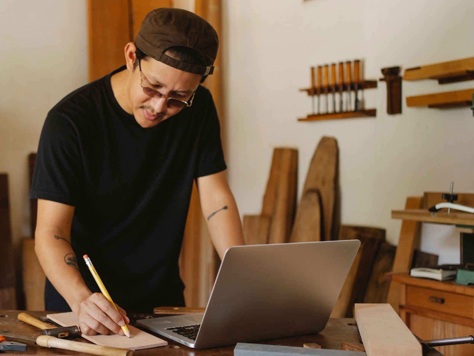 Furniture maker in his workshop writing measurements on a board by hand, with an open laptop next to him on the workbench