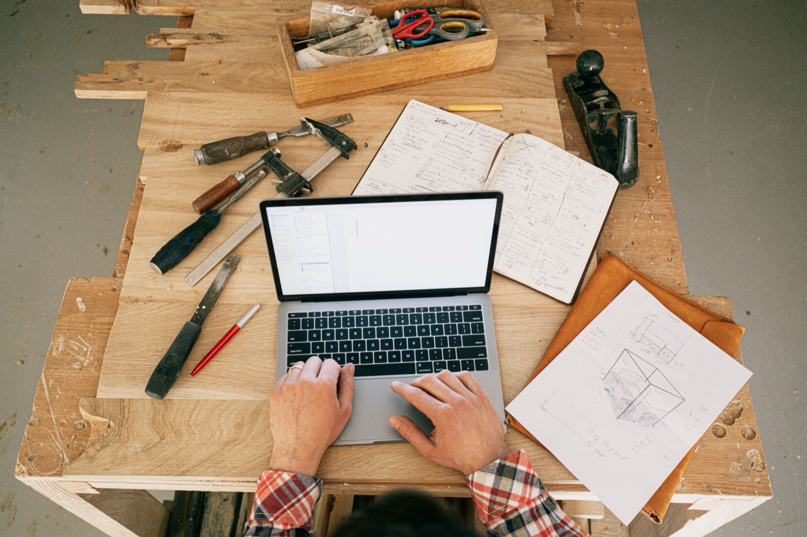 Overhead view of a workbench with a laptop, a handwritten notebook, a loose sketch on paper and tools lying around each other